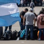 Bandeira da Argentina em destaque em primeiro plano, com grupo de turistas brasileiros desanimados e bagagens ao fundo, simbolizando o alto custo das viagens para a Argentina e o impacto econômico para os brasileiros.